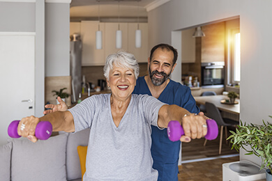Woman with white hair lifting lightweight dumbbells in her home with the guidance of a physical therapist.