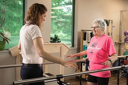 Older female patient in bright pink shirt using parallel bars to practice walking