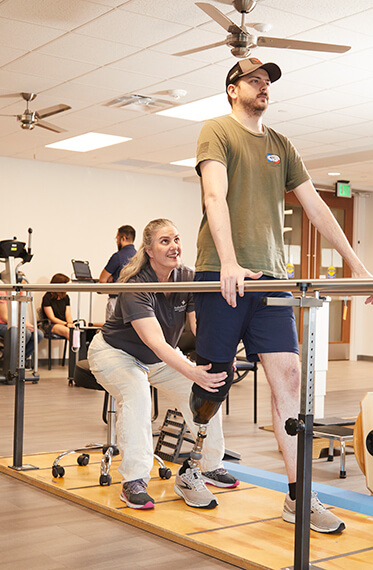 Photo of a man using balance bars to practice walking with a prosthetic leg. A therapist is behind him for support